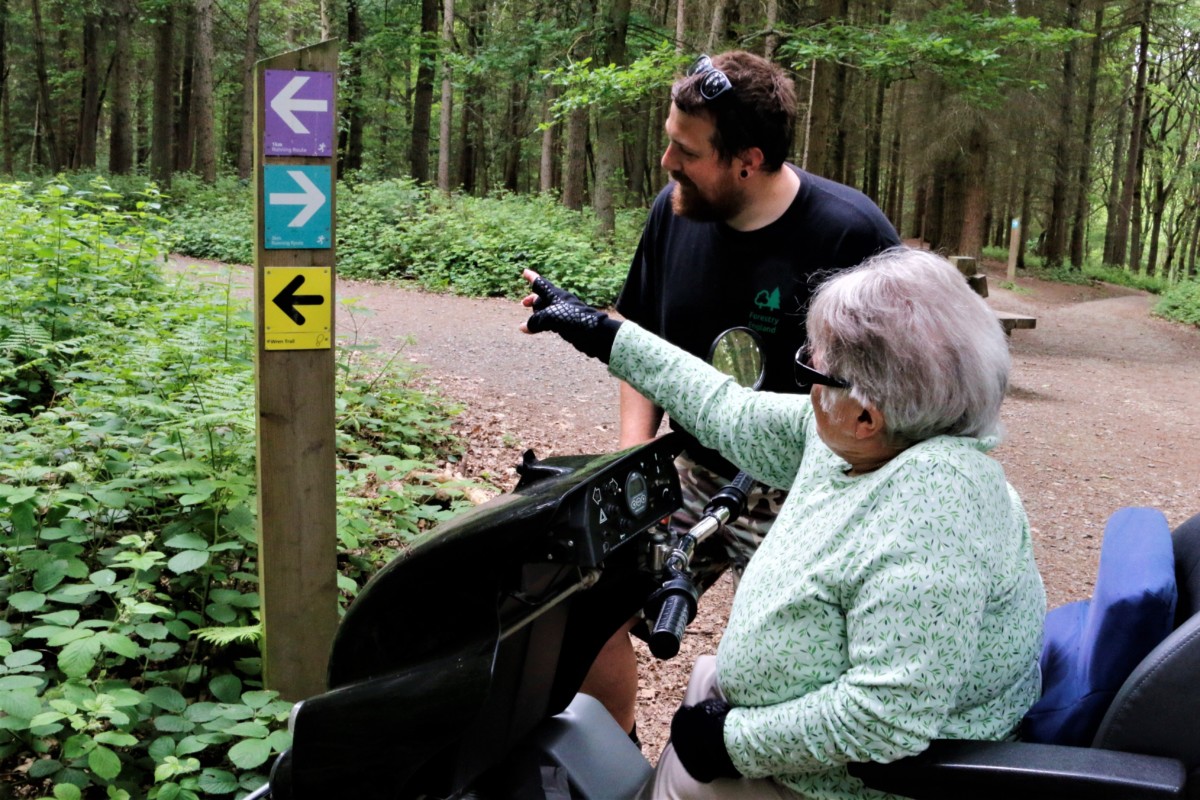 Living Options Devon volunteer undertaking a walk and talk audit with customer. They are discussing signage on a woodland gravel path.