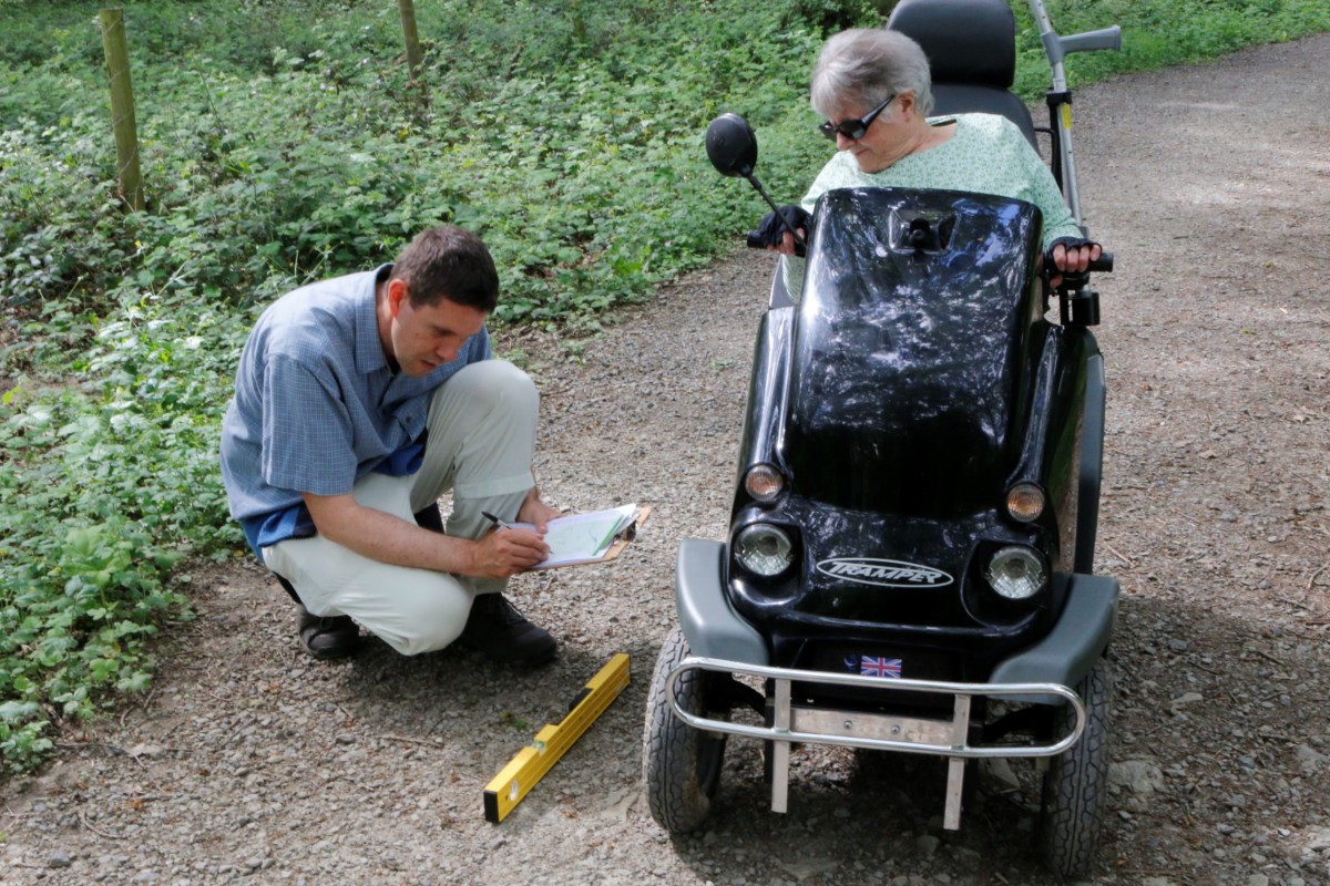 Neil undertaking a disabilioty access audit with a Living Options Devon volunteer, they are on a gravel part in the countryside. Neil has a tape measure and the volunteer is in al all terrain mobility scooter.